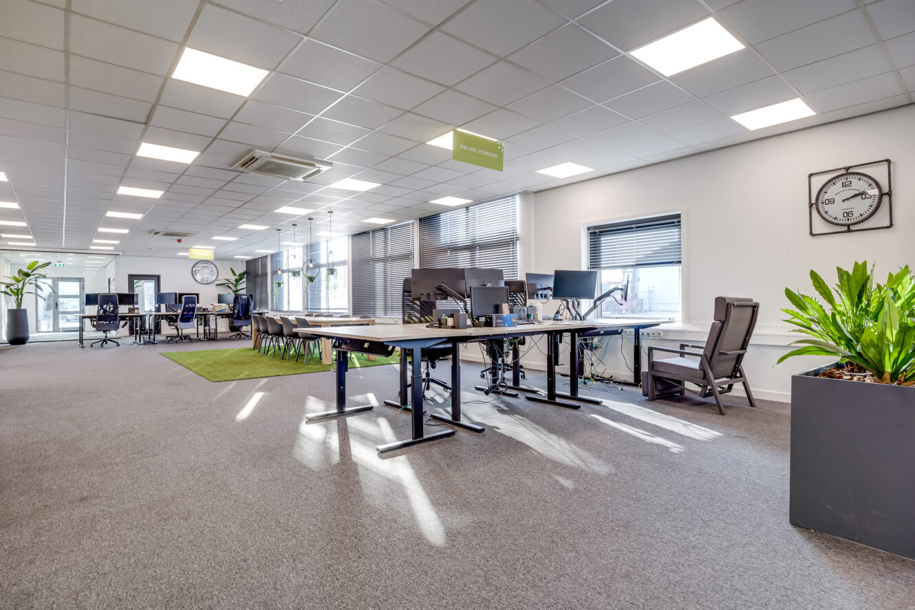 Modern open kantoor met verschillende lege bureaus, ergonomische stoelen, computers, grote ramen, kamerplanten en wandklokken. Heldere verlichting en een schone, ruime omgeving.