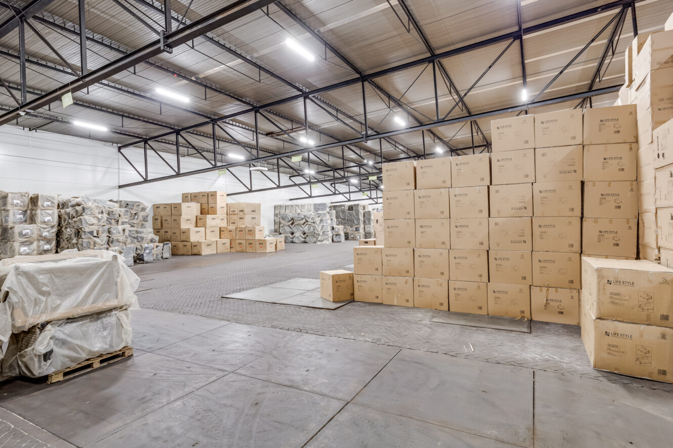 Magazijninterieur met gestapelde kartonnen dozen en ingepakte pallets, georganiseerd in rijen onder heldere industriële verlichting.