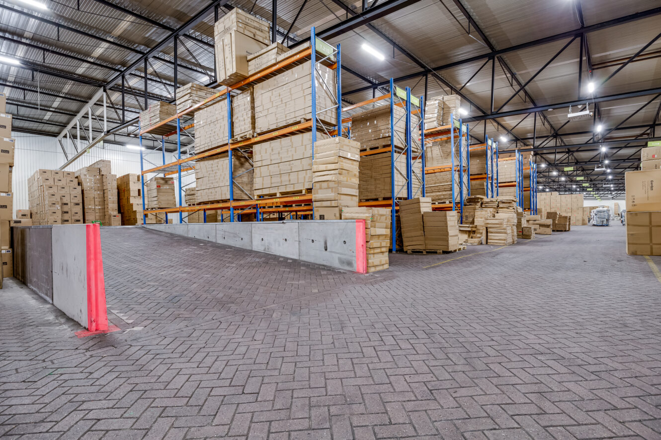 Interieur van een magazijn met hoge rekken gevuld met gestapelde kartonnen dozen en een betonnen helling die naar opslagruimtes leidt.