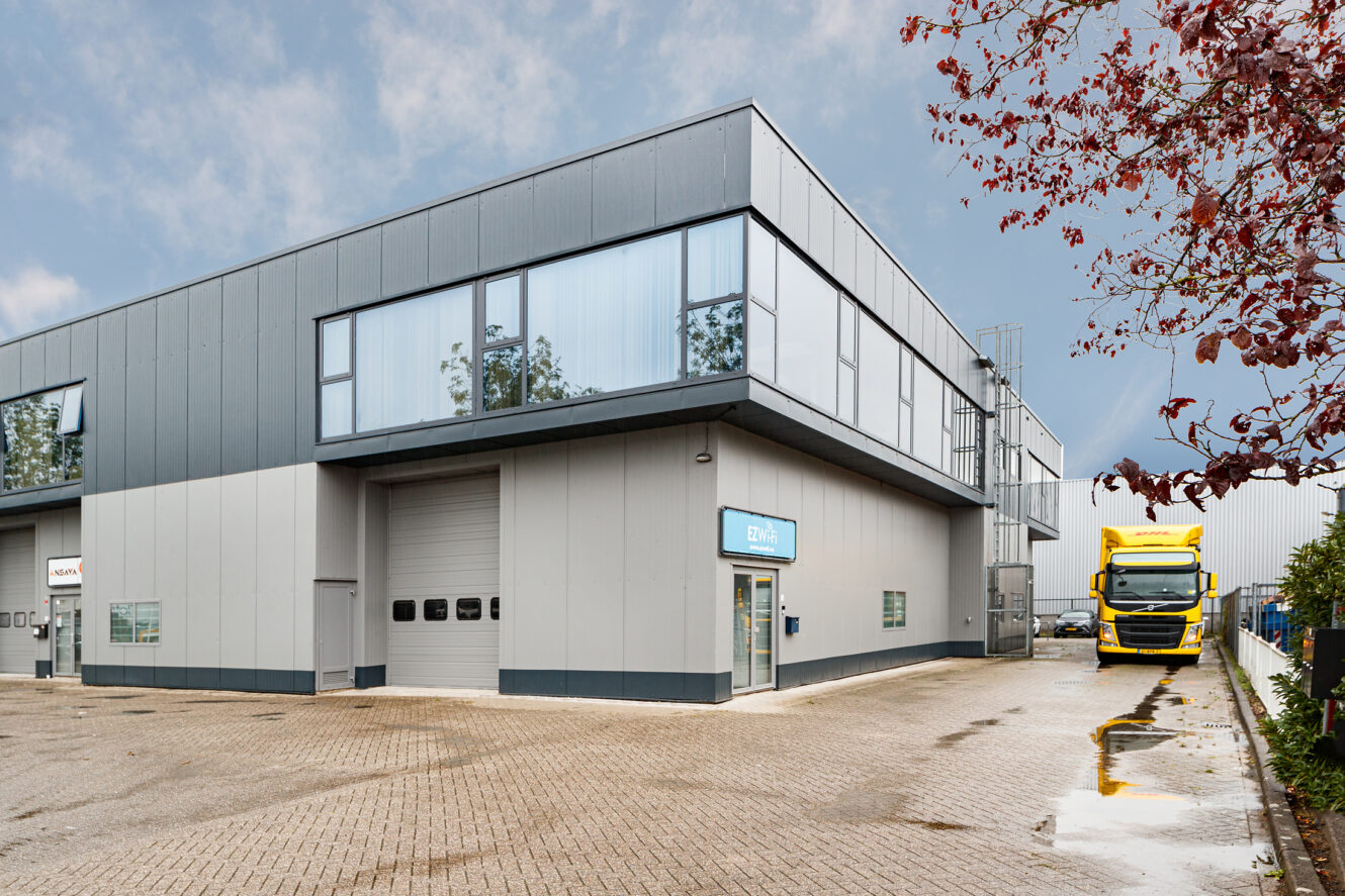 Een modern industrieel gebouw met grote ramen en een grijze buitenkant staat naast een geparkeerde gele vrachtwagen onder een bewolkte hemel.