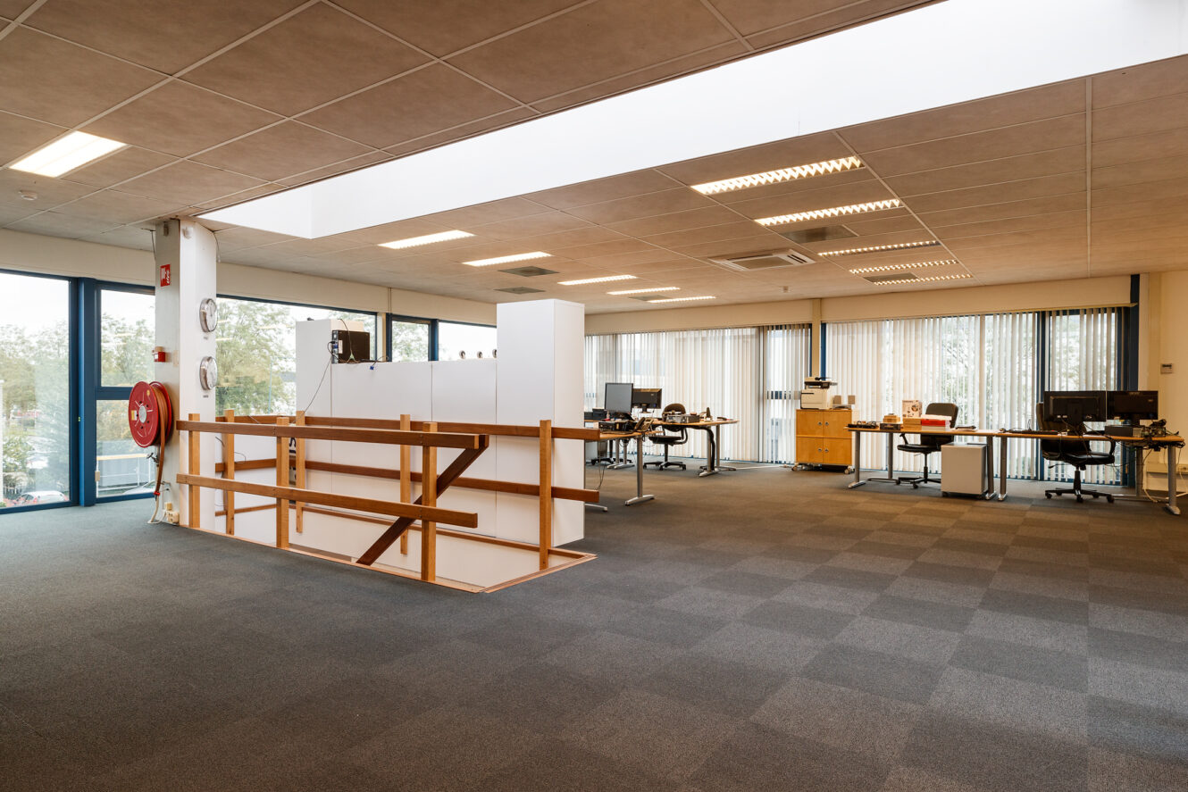 Ruim open kantoor met bureaus, computers, grote ramen en neutrale vloerbedekking, met minimaal meubilair en natuurlijk licht.
