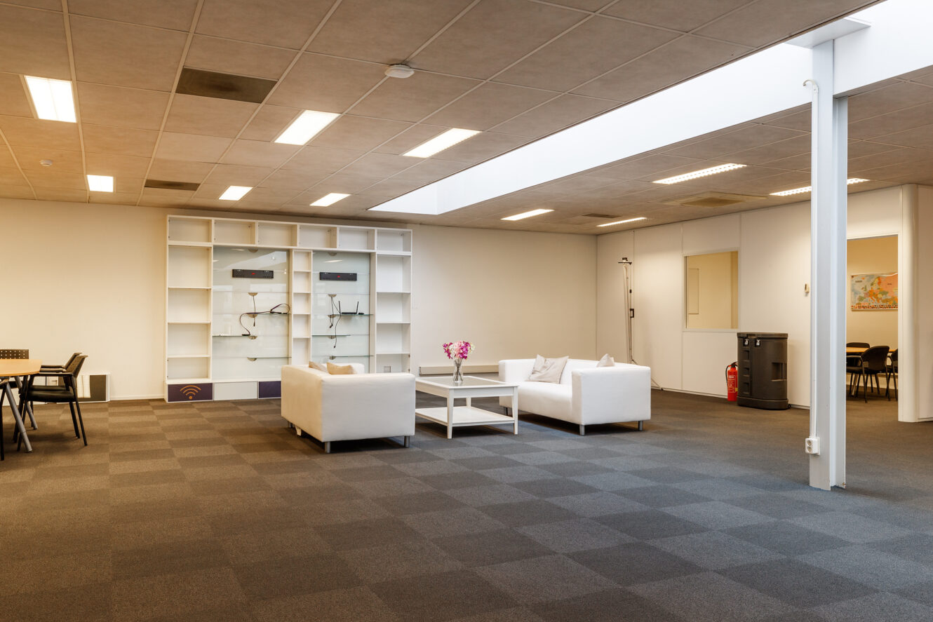 Moderne kantoorlounge met witte banken, een salontafel, planken en bovenverlichting. Er zijn stoelen en tafels zichtbaar in aangrenzende kamers door open deuropeningen.