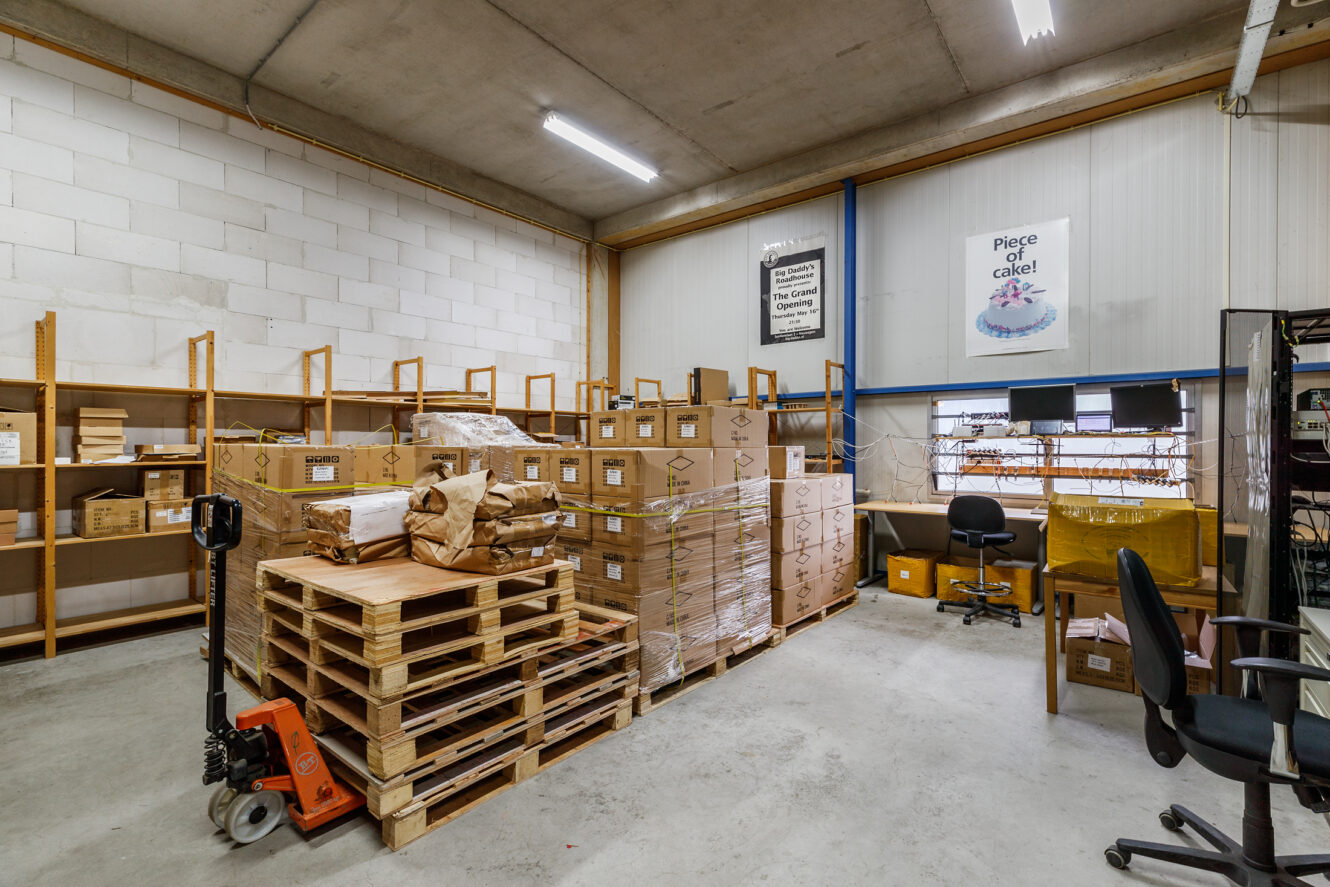 Een magazijnruimte met gestapelde kartonnen dozen op pallets, lege houten planken, een palletkrik, twee bureaustoelen, bureaus, computers en motiverende posters aan de muur.