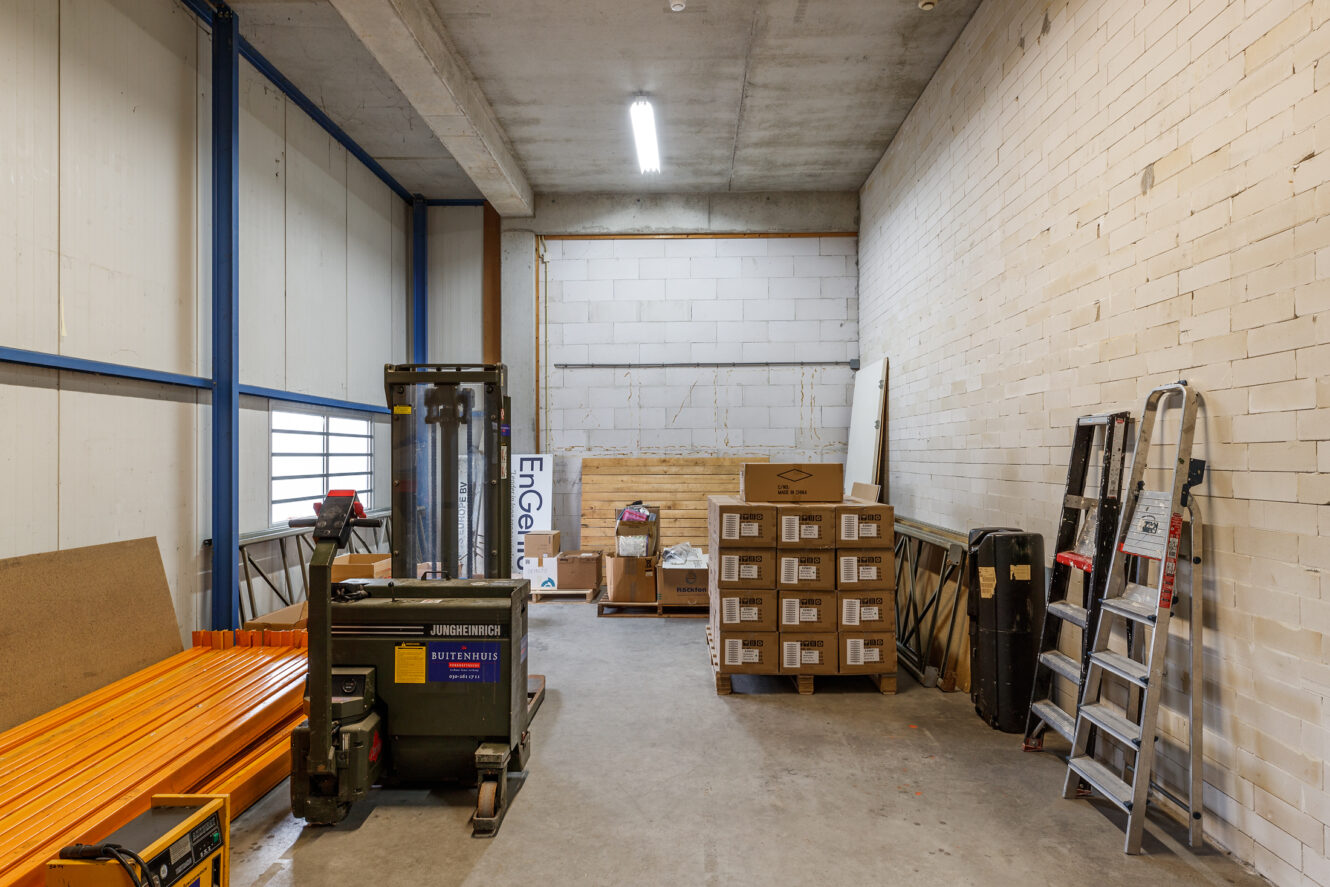 Opslagruimte met gestapelde dozen, een vorkheftruck, oranje pijpen, ladders en diverse voorwerpen tegen witte bakstenen muren en een betonnen plafond.