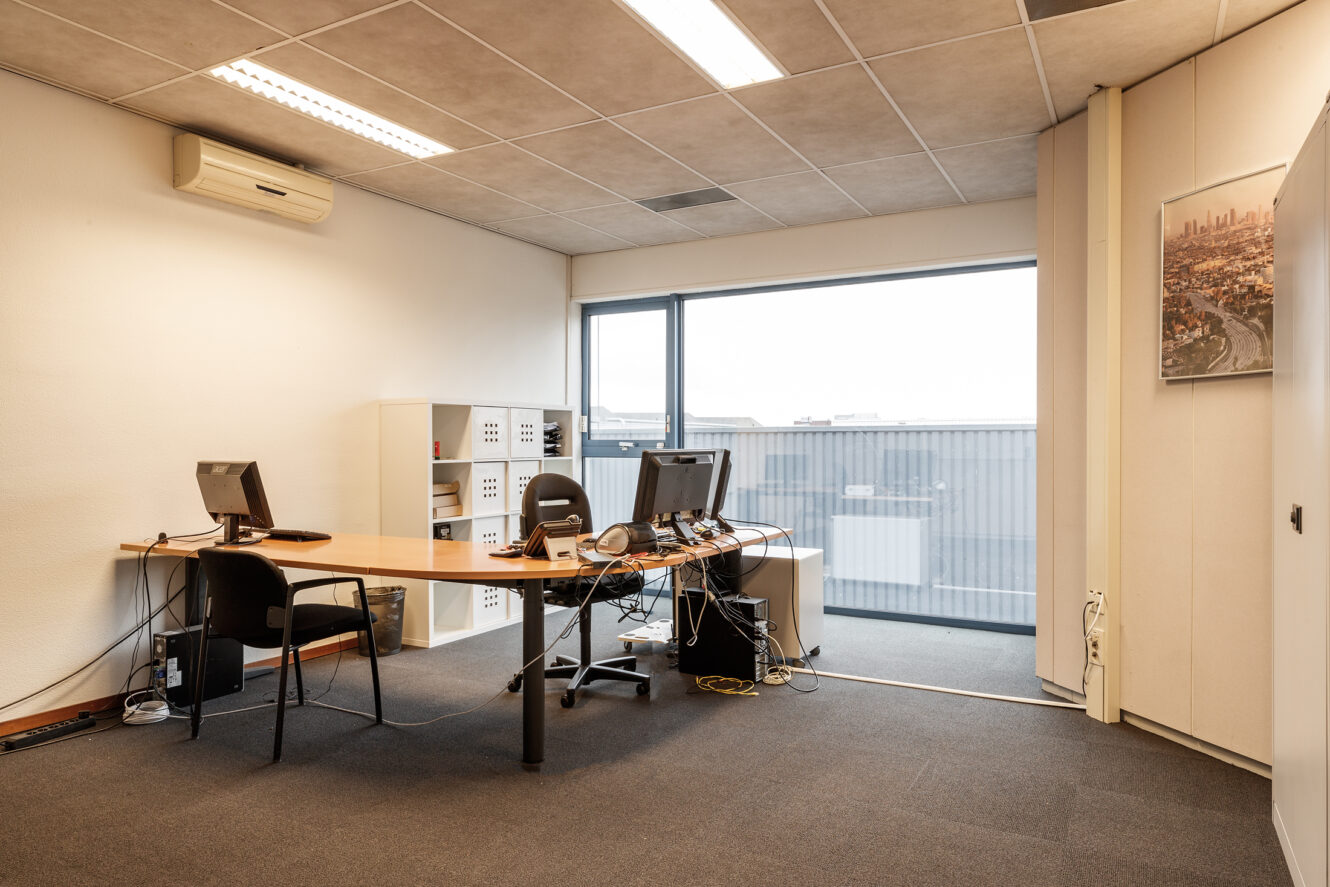 Een klein kantoor met twee bureaus, computers, bureaustoelen, planken en grote ramen met uitzicht op een industriegebied.