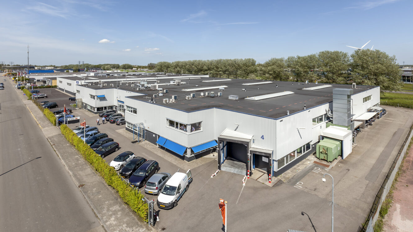 Een groot industrieel pakhuis met een parkeerplaats, verschillende auto's buiten geparkeerd en laadkades, gelegen langs een brede straat onder een heldere hemel.