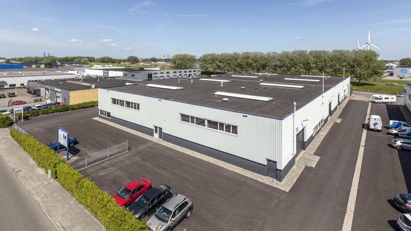 Een groot industrieel pakhuis met een plat dak wordt omringd door verharde wegen en geparkeerde auto's, met andere commerciële gebouwen en bomen op de achtergrond.