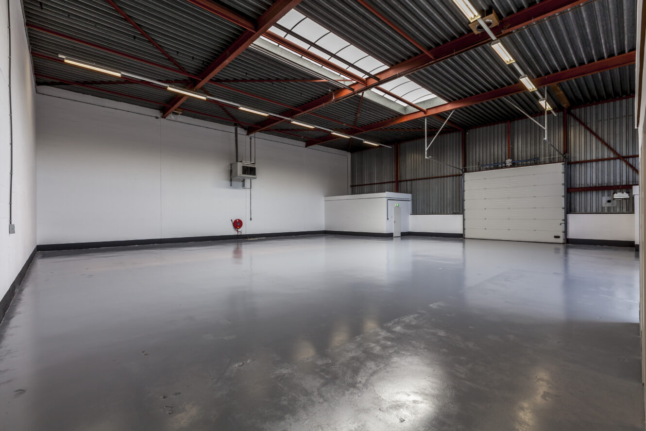 Leeg industrieel magazijn met betonnen vloeren, witte muren, hoge plafonds, zichtbare balken en een kleine afgesloten kantoorruimte in de hoek. Rechts is een grote roldeur zichtbaar.