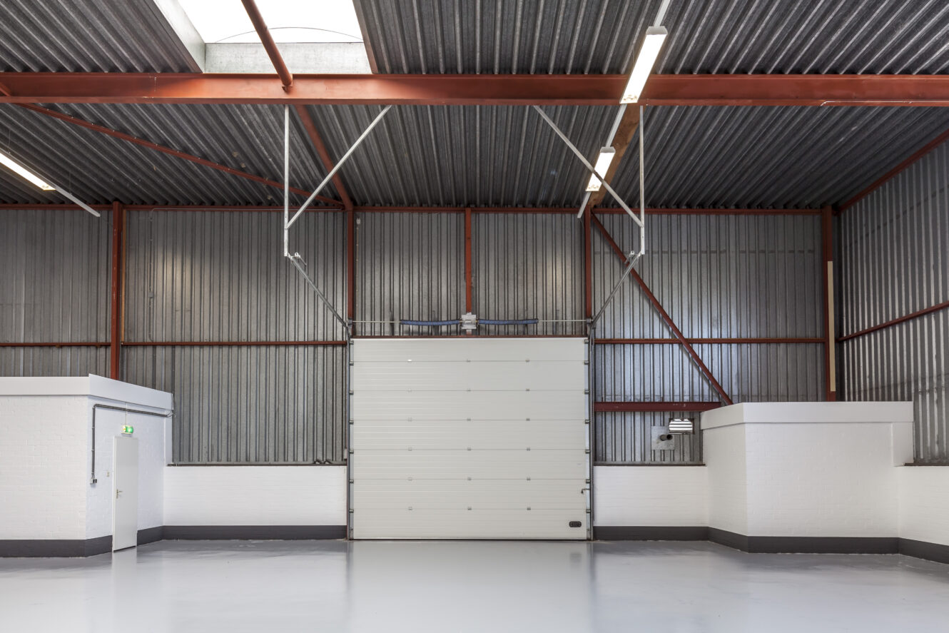 Interieur van een leeg industrieel pakhuis met een grote gesloten overheaddeur, witte muren en zichtbare metalen balken en plafond.