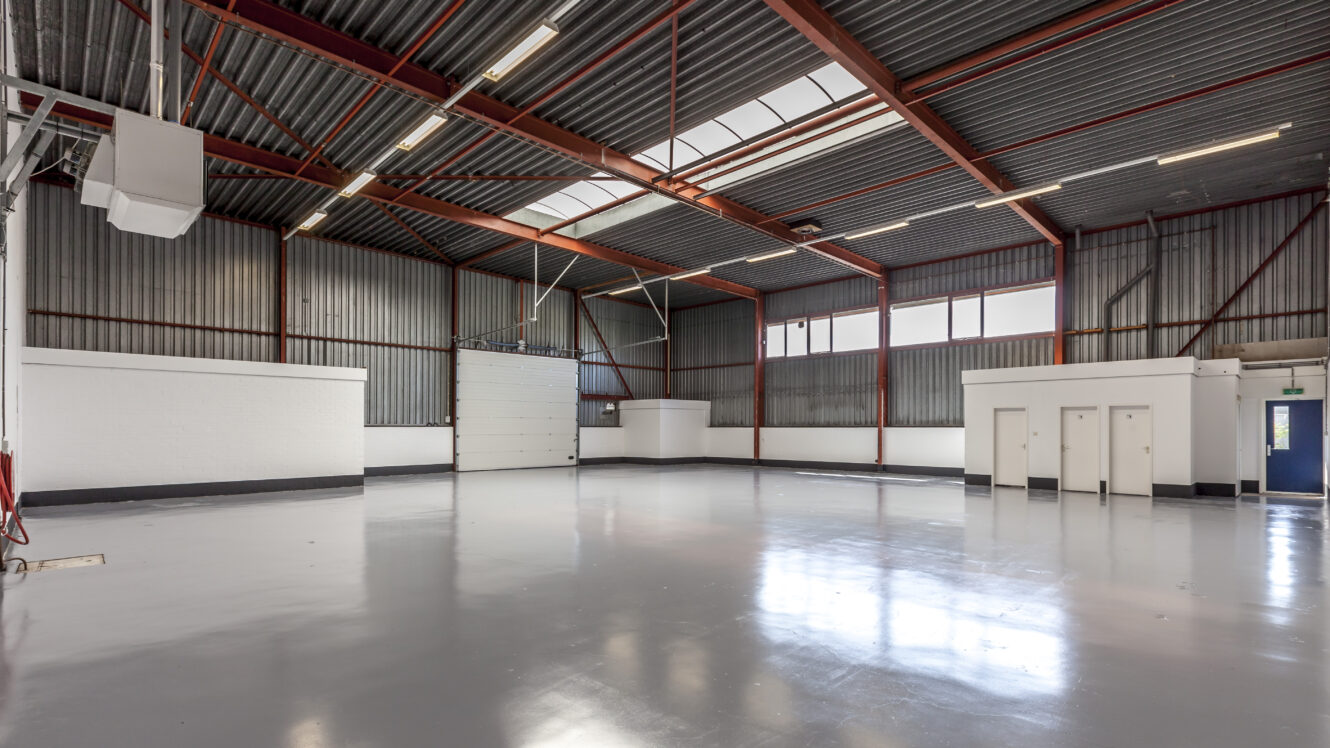 Leegstaand industrieel pakhuis met gepolijste betonnen vloer, rode stalen balken, witte scheidingswanden, hoog plafond en grote ramen die natuurlijk licht binnenlaten.