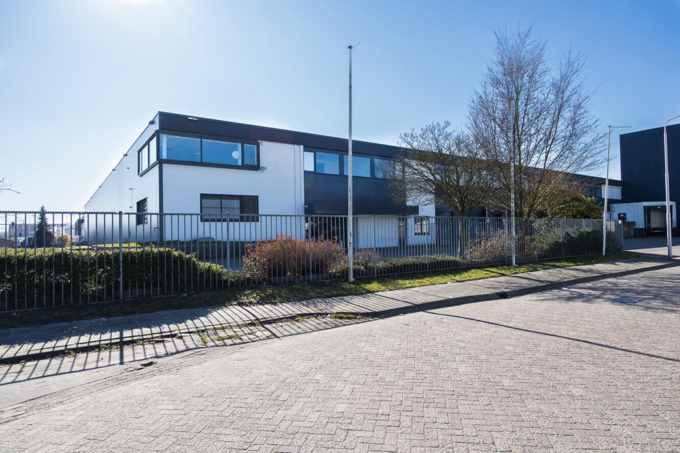 Een groot modern industrieel gebouw met witte en donkergrijze buitenkant, omringd door een metalen hek en tuinarchitectuur, op een zonnige dag.