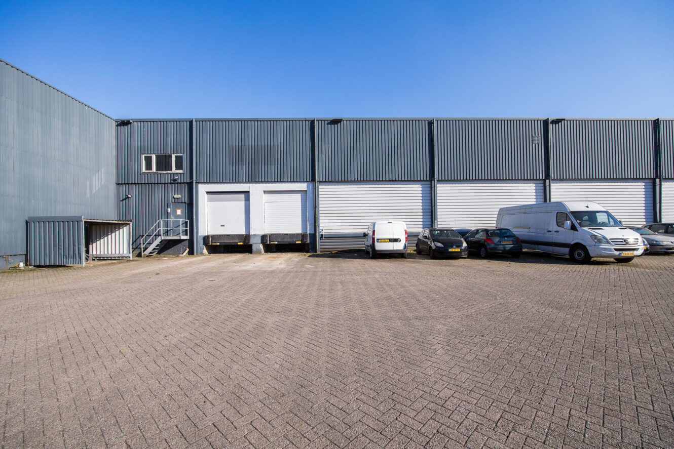 Een groot industrieel magazijn met laadkades en verschillende geparkeerde voertuigen, waaronder bestelwagens en auto's, op een verhard terrein onder een strakblauwe hemel.