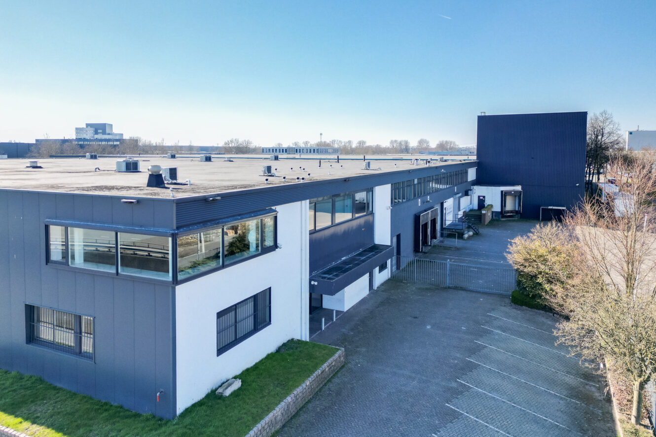 Een modern industrieel of kantoorgebouw met een donkerblauwe en witte buitenkant, grote ramen, een geplaveide parkeerplaats en een strakblauwe lucht.