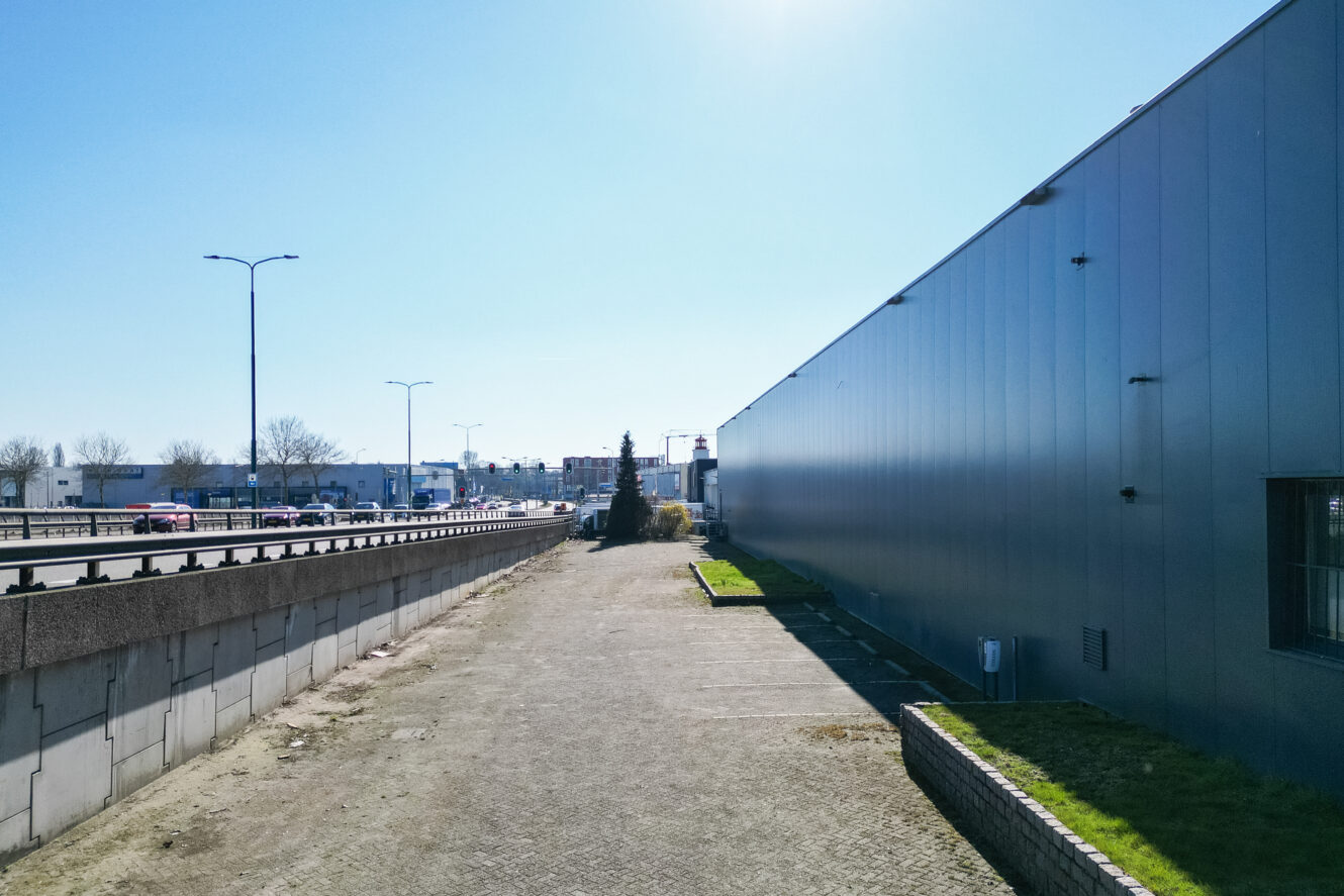 Een brede stoep loopt langs een grijs industrieel gebouw, met links een weg en verkeer en daarboven een strakblauwe lucht.
