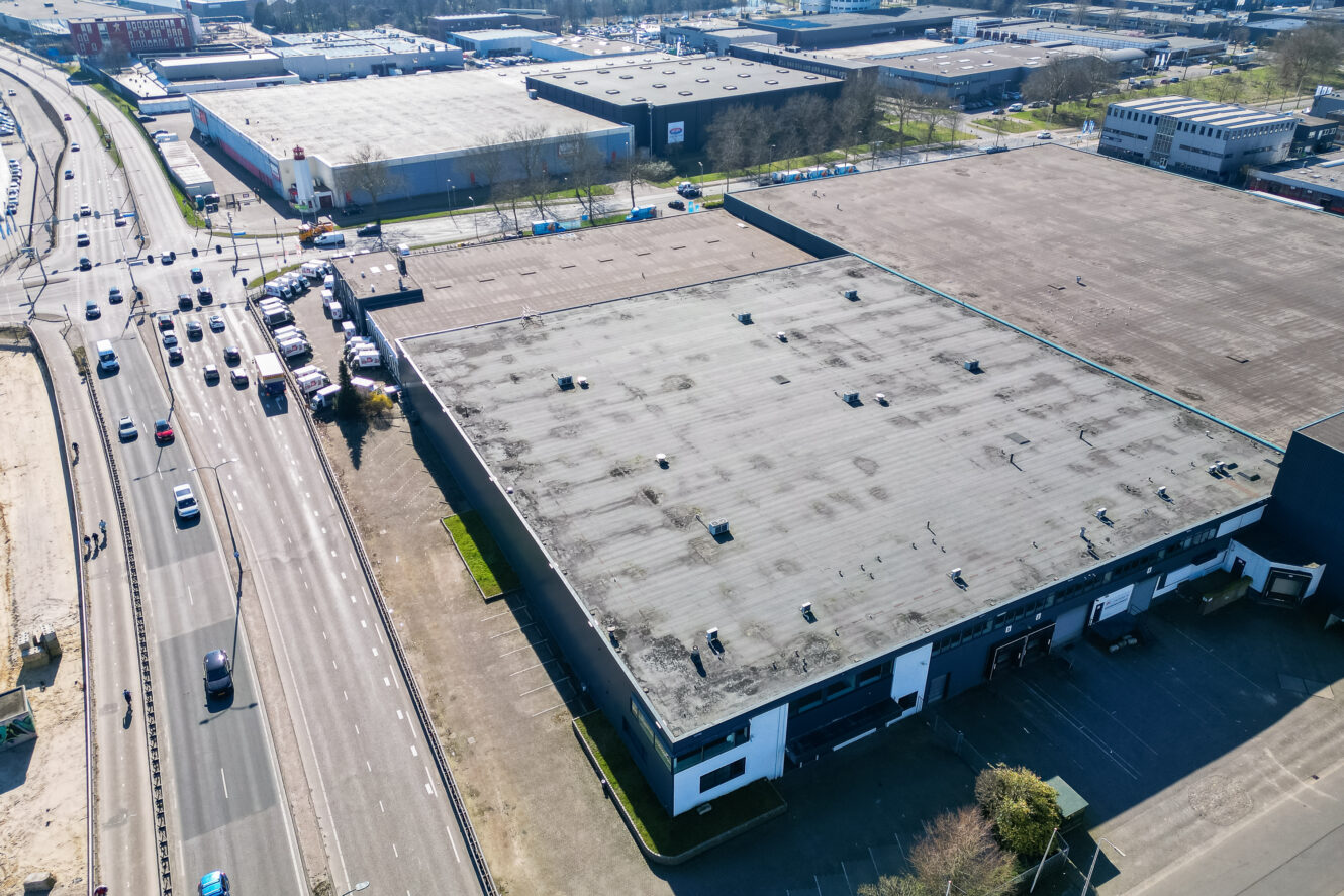 Luchtfoto van een groot industrieel pakhuis met een plat dak, naast een drukke weg en parkeerplaats, in een stedelijk industriegebied.
