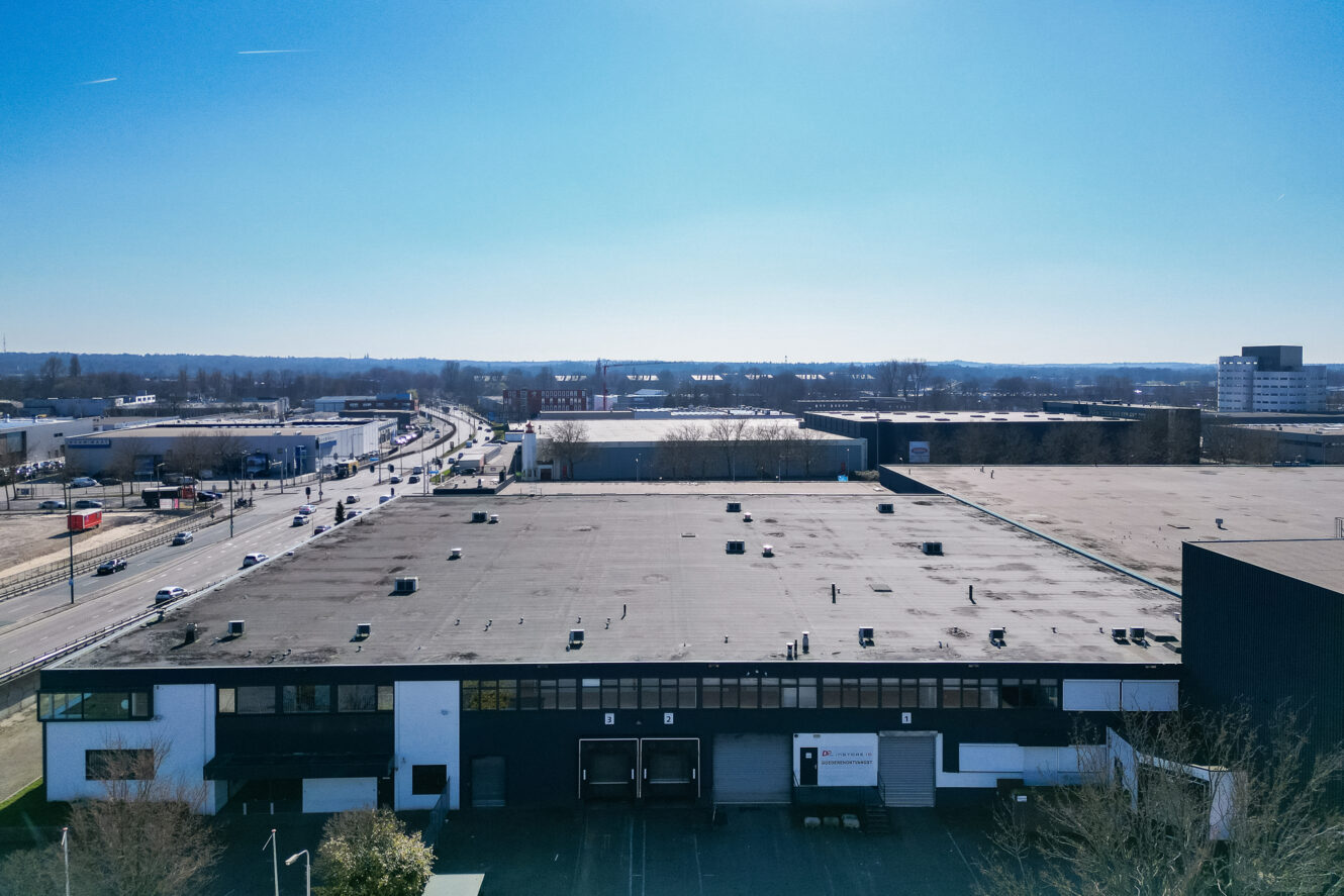Luchtfoto van een groot industrieel gebouw met platte daken naast een weg, met andere magazijnen en een heldere blauwe lucht op de achtergrond.