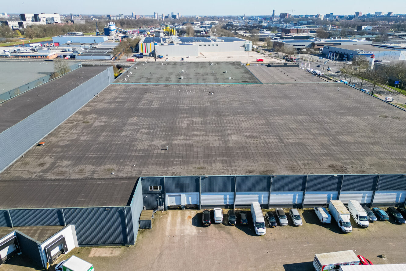 Luchtfoto van een groot industrieel pakhuis met een plat dak, vrachtwagens geparkeerd bij laadkades en omliggende stedelijke gebouwen.
