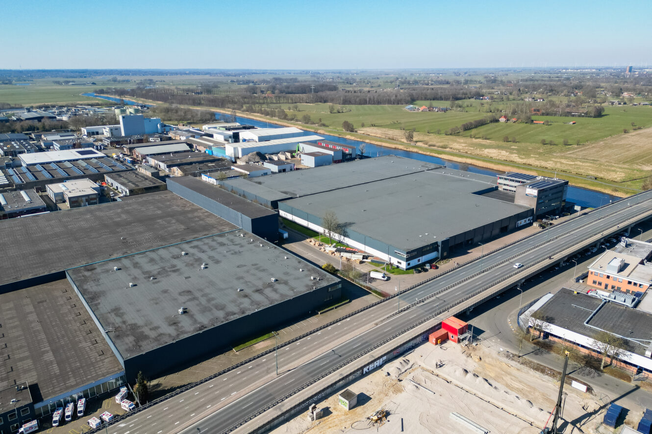 Luchtfoto van een industriegebied met grote magazijnen, een weg en aangrenzende groene velden onder een heldere hemel.