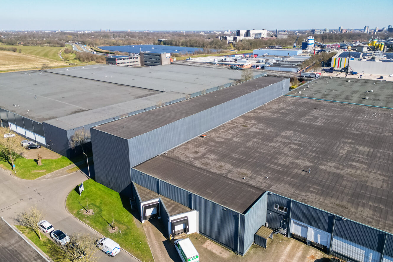 Luchtfoto van grote industriële magazijngebouwen met laadkades, omgeven door wegen, gras en geparkeerde voertuigen.
