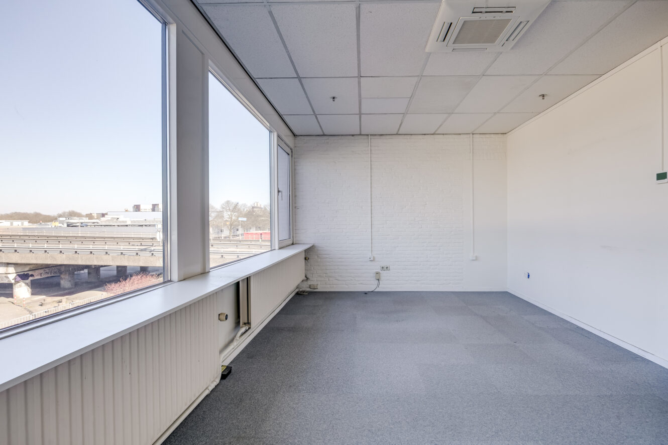 Lege kantoorruimte met grote ramen, witte muren, grijs tapijt en plafondpanelen; uitzicht op een treinspoor en gebouwen buiten.