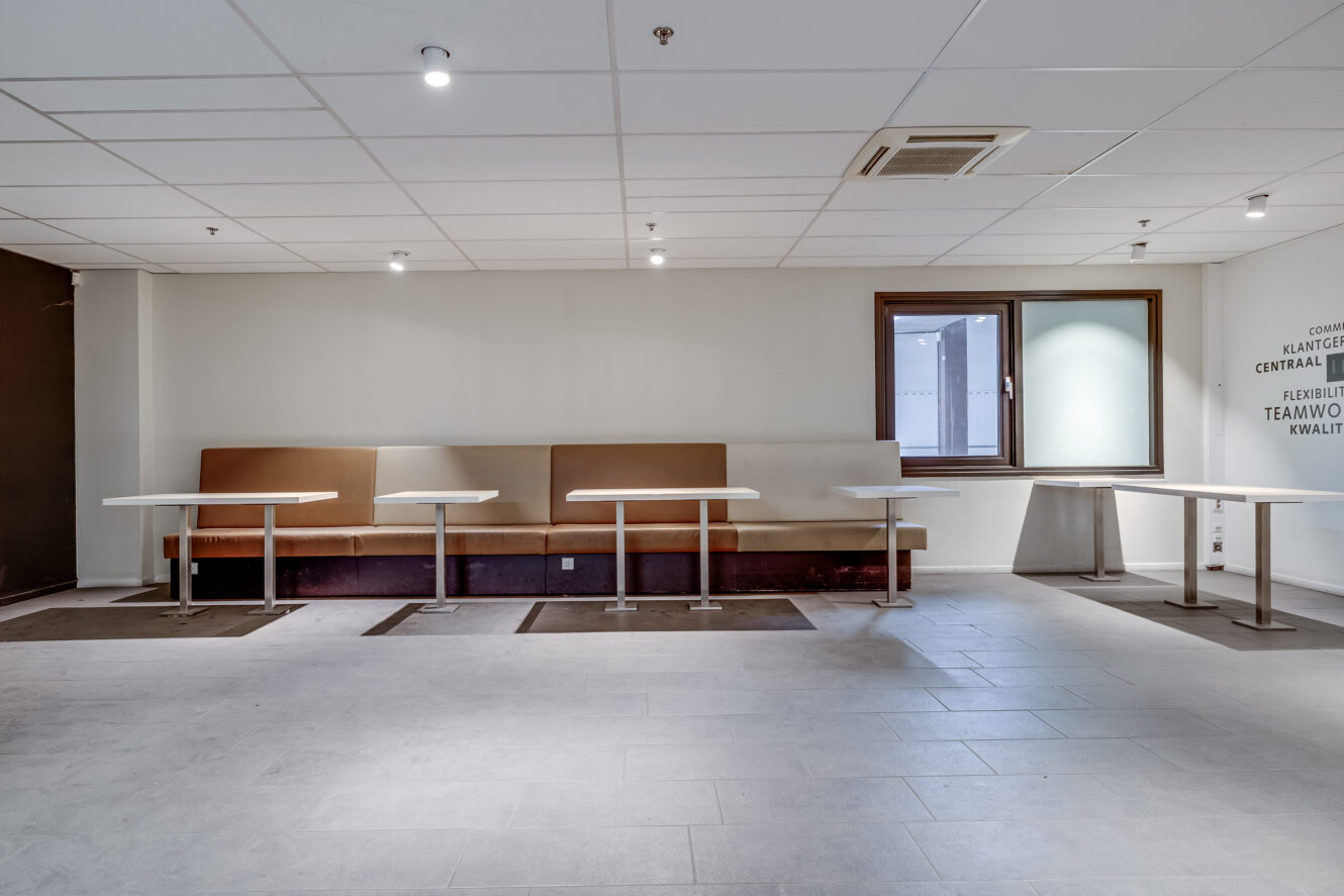 Lege cafetaria met bruine zitbanken, witte tafels, tegelvloer, wit plafond en een klein raam aan de rechtermuur.