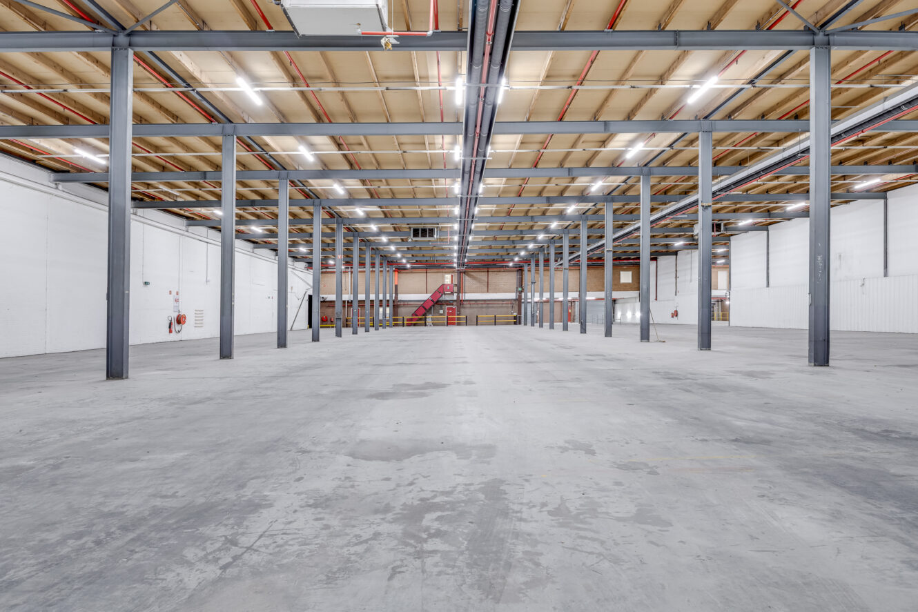 Interieur van een leeg magazijn met betonnen vloer, zichtbare metalen balken, hoog plafond en tl-verlichting. Rode trappen en brandblussers zijn zichtbaar aan het uiteinde.