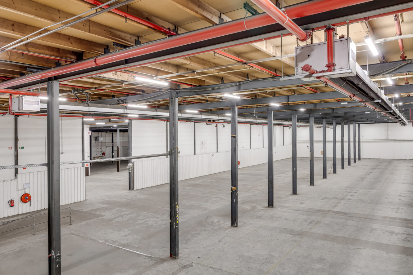 Interieur van een leeg pakhuis met betonnen vloeren, metalen steunpilaren, witte scheidingswanden en zichtbare plafondpijpen. Heldere bovenverlichting verlicht de ruimte.