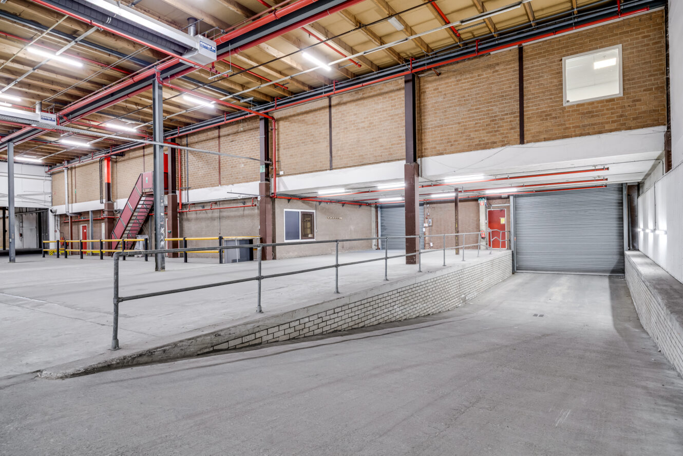 Interieur van een leeg industrieel pakhuis met een betonnen helling, metalen leuningen, hoog plafond, zichtbare buizen, bakstenen muren en een opgerolde garagedeur.