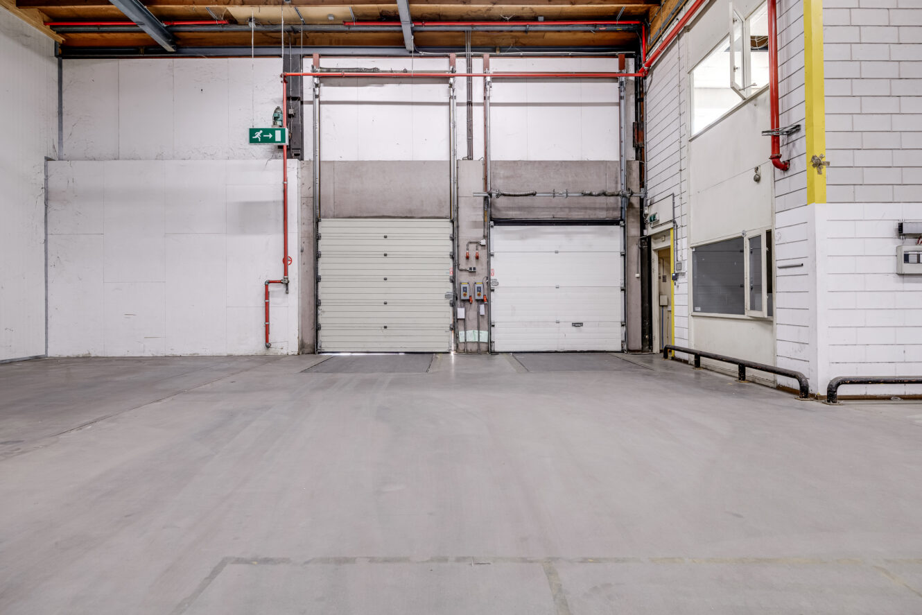 Leeg industrieel magazijninterieur met twee gesloten laadperrondeuren, zichtbare plafonds, betonnen vloer en zichtbare leidingen en elektrische panelen.