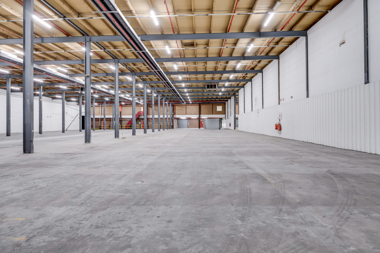 Interieur van een leeg industrieel pakhuis met betonnen vloer, zichtbare balken, witte muren en bovenverlichting.