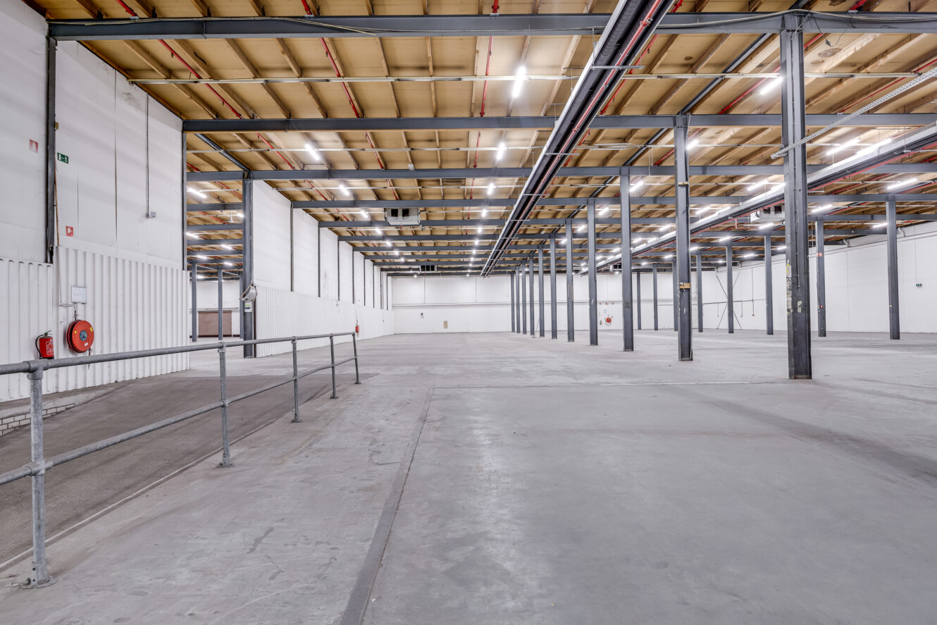 Leeg industrieel magazijninterieur met hoge plafonds, zichtbare balken, metalen steunpilaren, witte muren en betonnen vloeren. Een metalen reling loopt langs een helling aan de linkerkant.