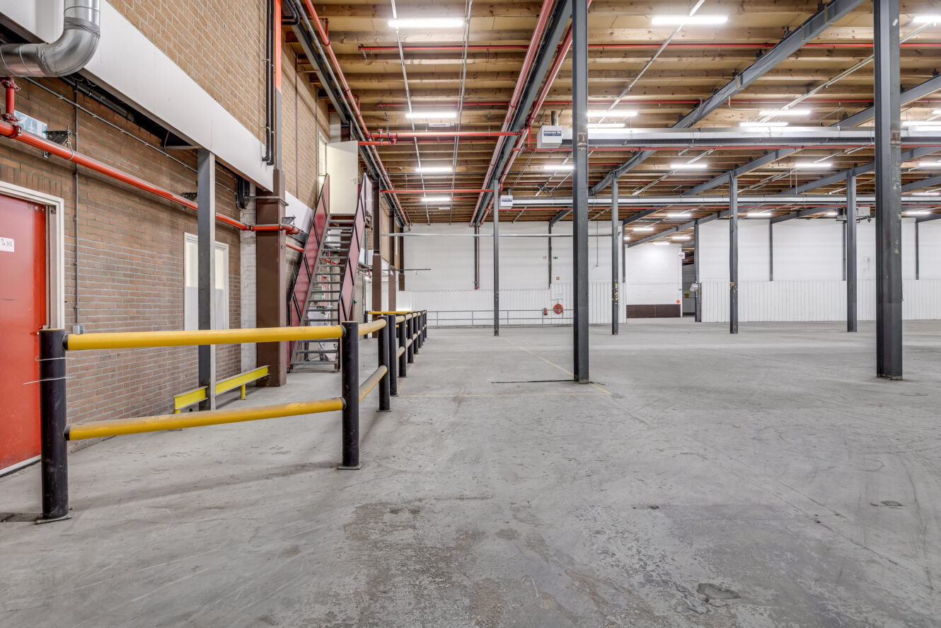 Interieur van een leeg industrieel pakhuis met een opengewerkt plafond, steunpilaren, een rode deur, gele veiligheidsleuningen en een metalen trap.