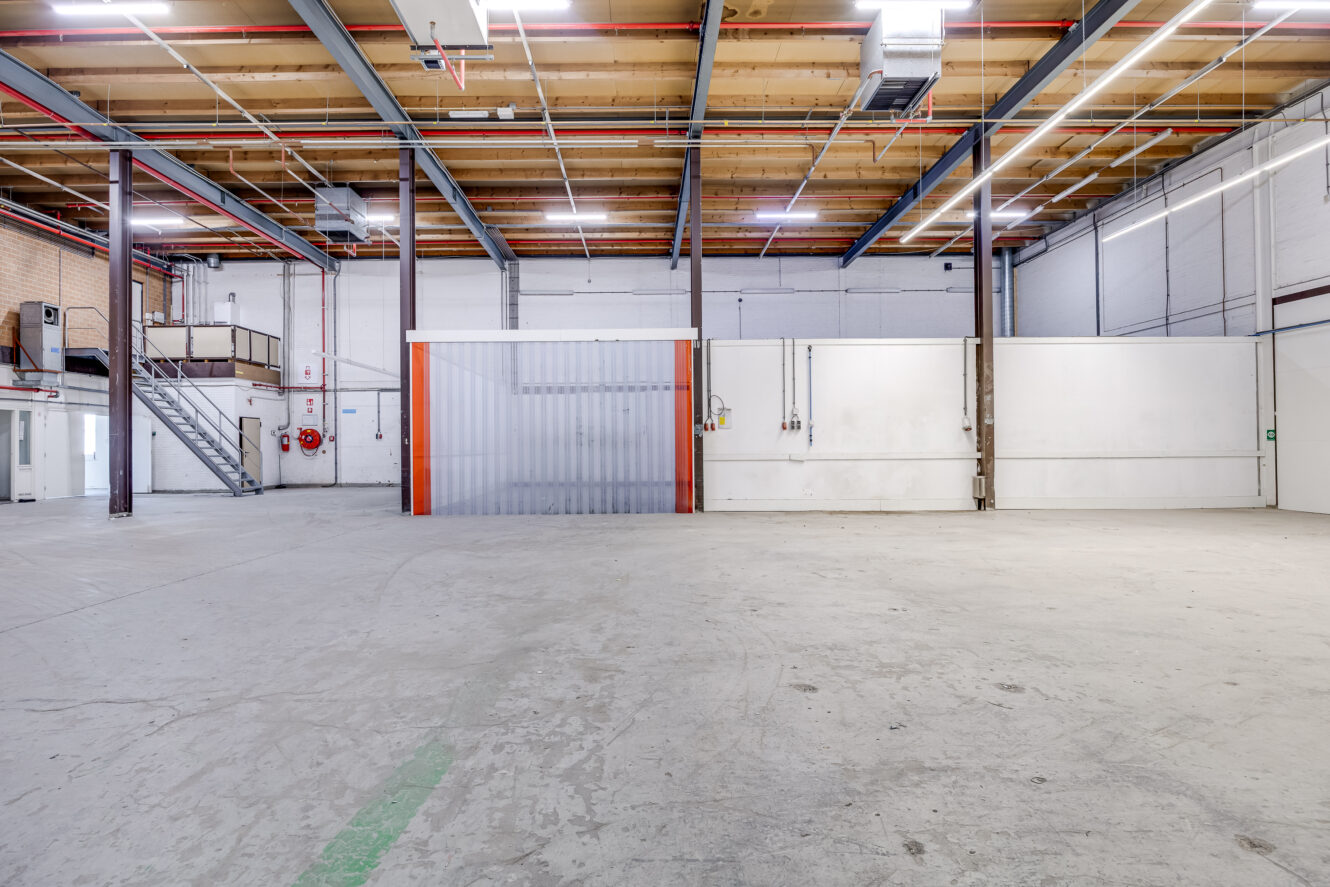 Interieur van een leeg industrieel pakhuis met hoge plafonds, zichtbare balken, een grote zeecontainer en een tussenverdieping aan de linkerkant.