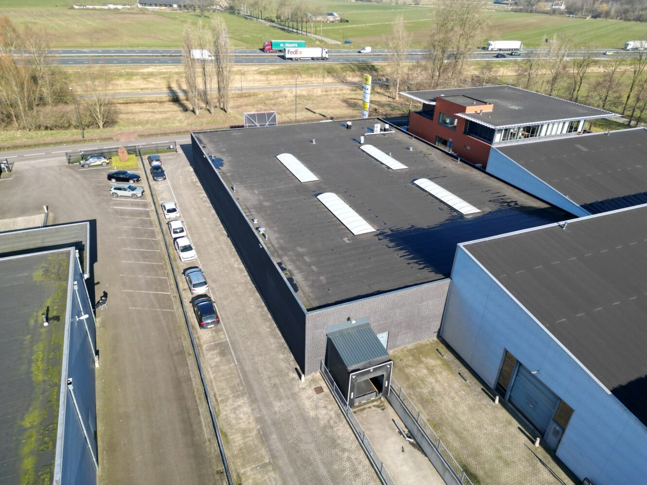 Luchtfoto van een industrieel gebouw met een plat dak, aangrenzende parkeerplaatsen en nabijgelegen wegen met auto's en vrachtwagens.
