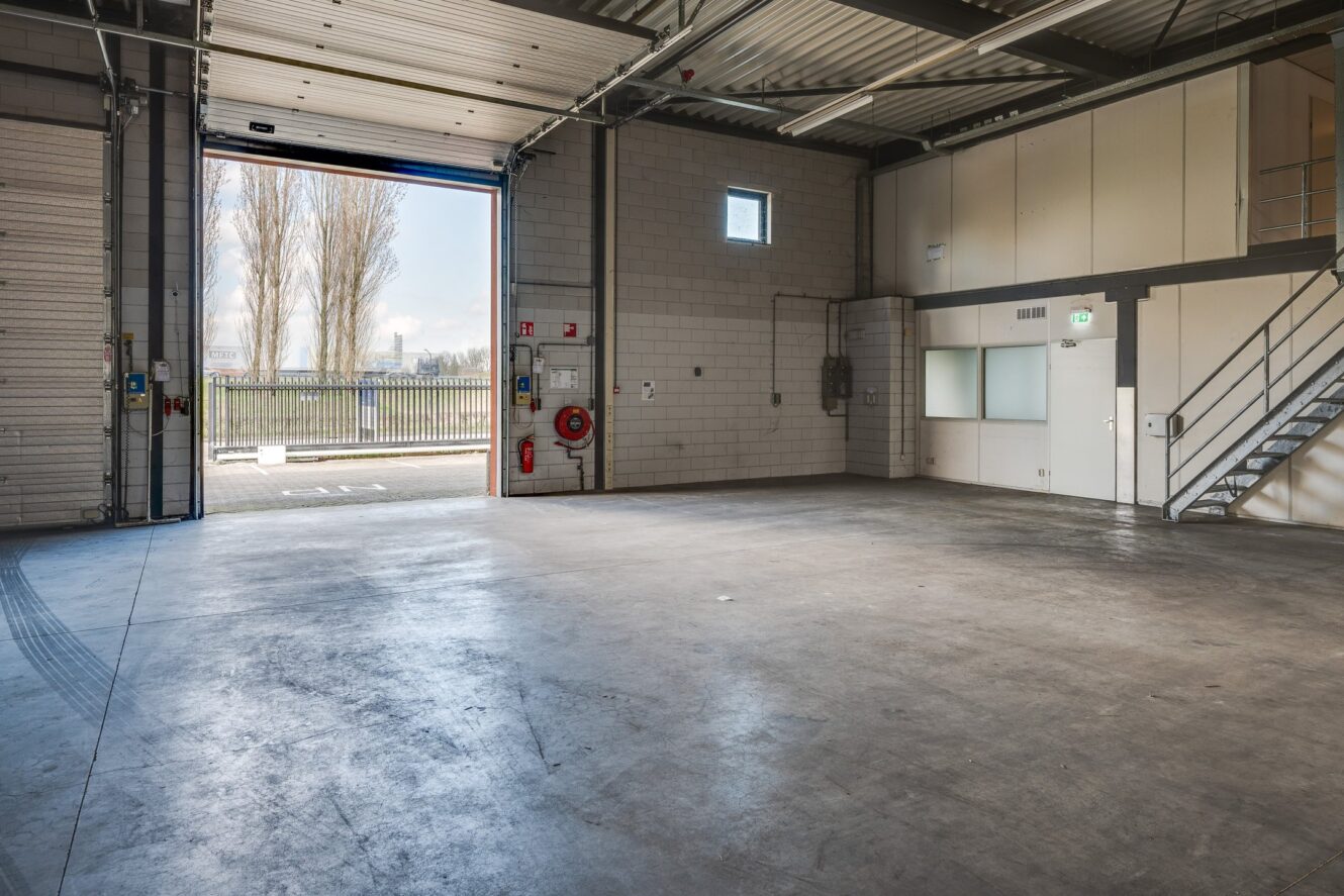 Een leeg industrieel pakhuis met betonnen vloer, hoog plafond, open garagedeur, trap, kleine ramen en zichtbare pijpen. Daglicht komt van buiten naar binnen.