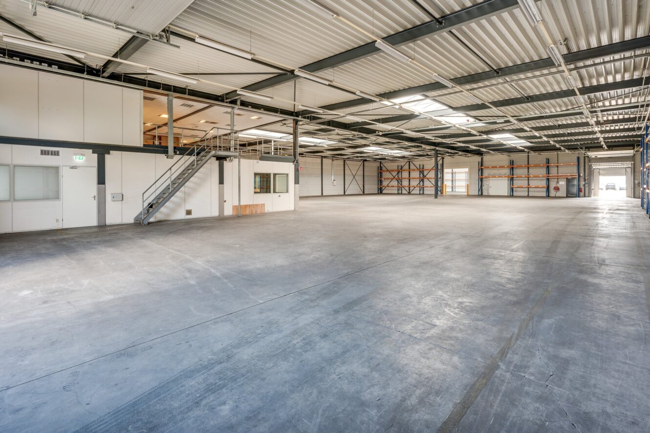 Groot leeg magazijninterieur met betonnen vloer, metalen plafondbalken, industriële verlichting, opslagrekken en een klein kantoorgedeelte met een trap naar een tussenverdieping.