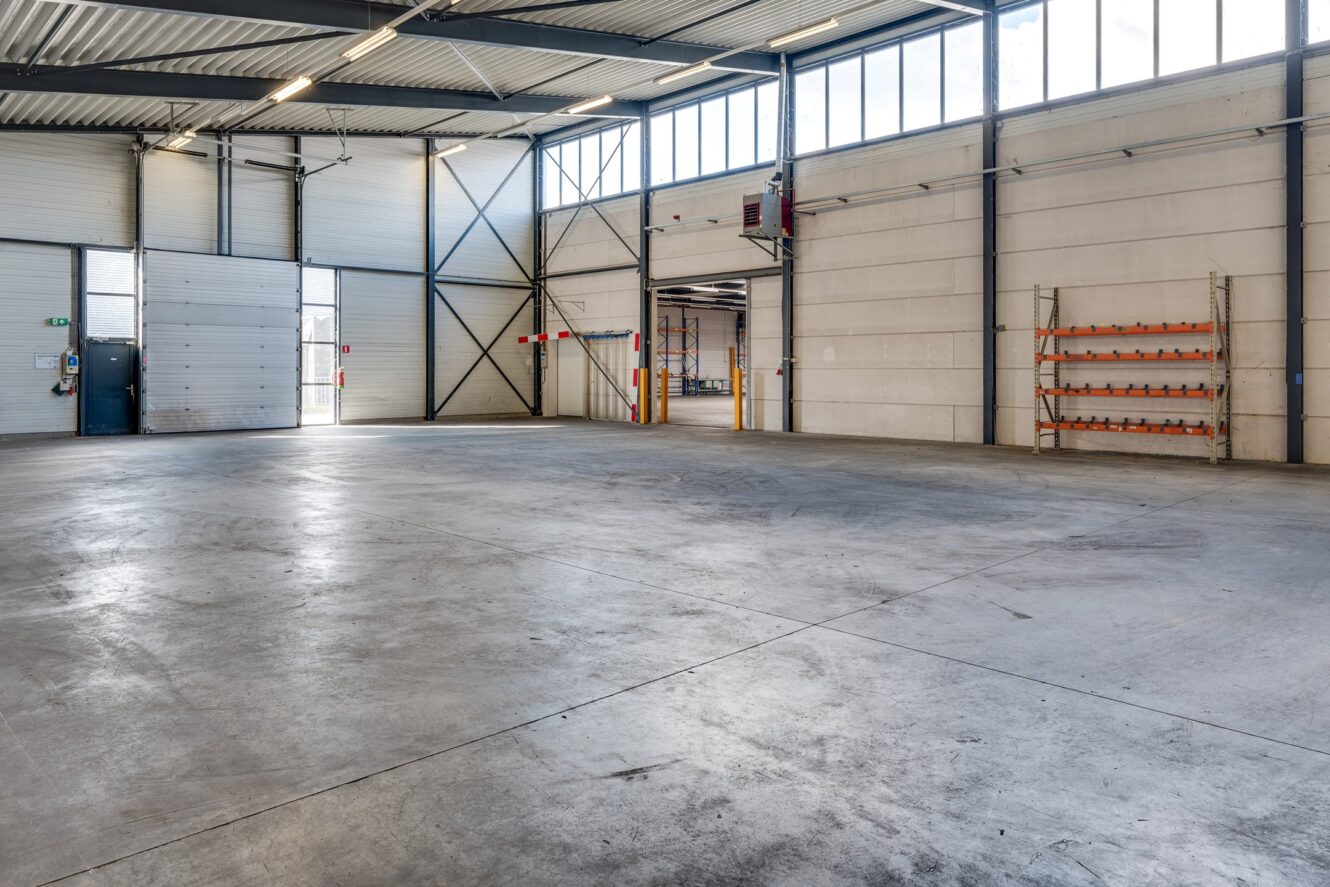 Interieur van een leeg industrieel magazijn met betonnen vloer, hoog plafond, grote ramen en overheaddeuren. Langs de muren zijn rekken en veiligheidspaaltjes zichtbaar.