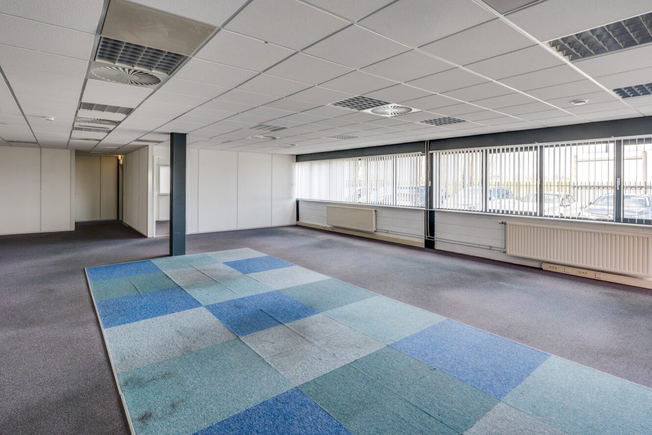 Een grotendeels lege kantoorruimte met blauwe en grijze tapijttegels, grote ramen met luxaflex en een verlaagd plafond met tl-verlichting.