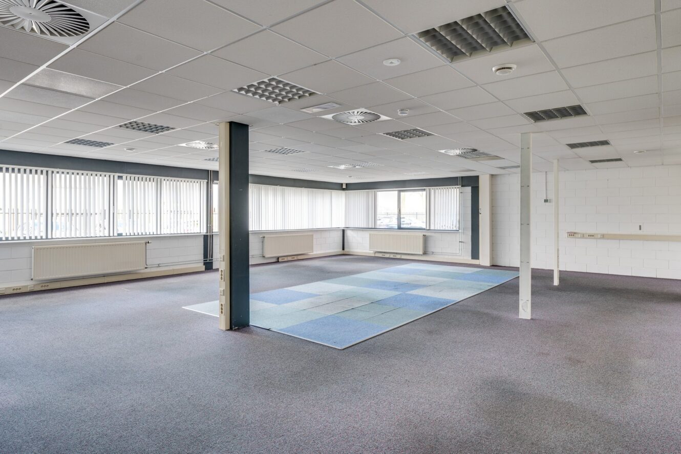 Grote, meestal lege kantoorkamer met grijs tapijt, een blauw vloerkleed, witte muren, verticale jaloezieën voor de ramen en een paar steunpalen en plafondlampen zichtbaar.