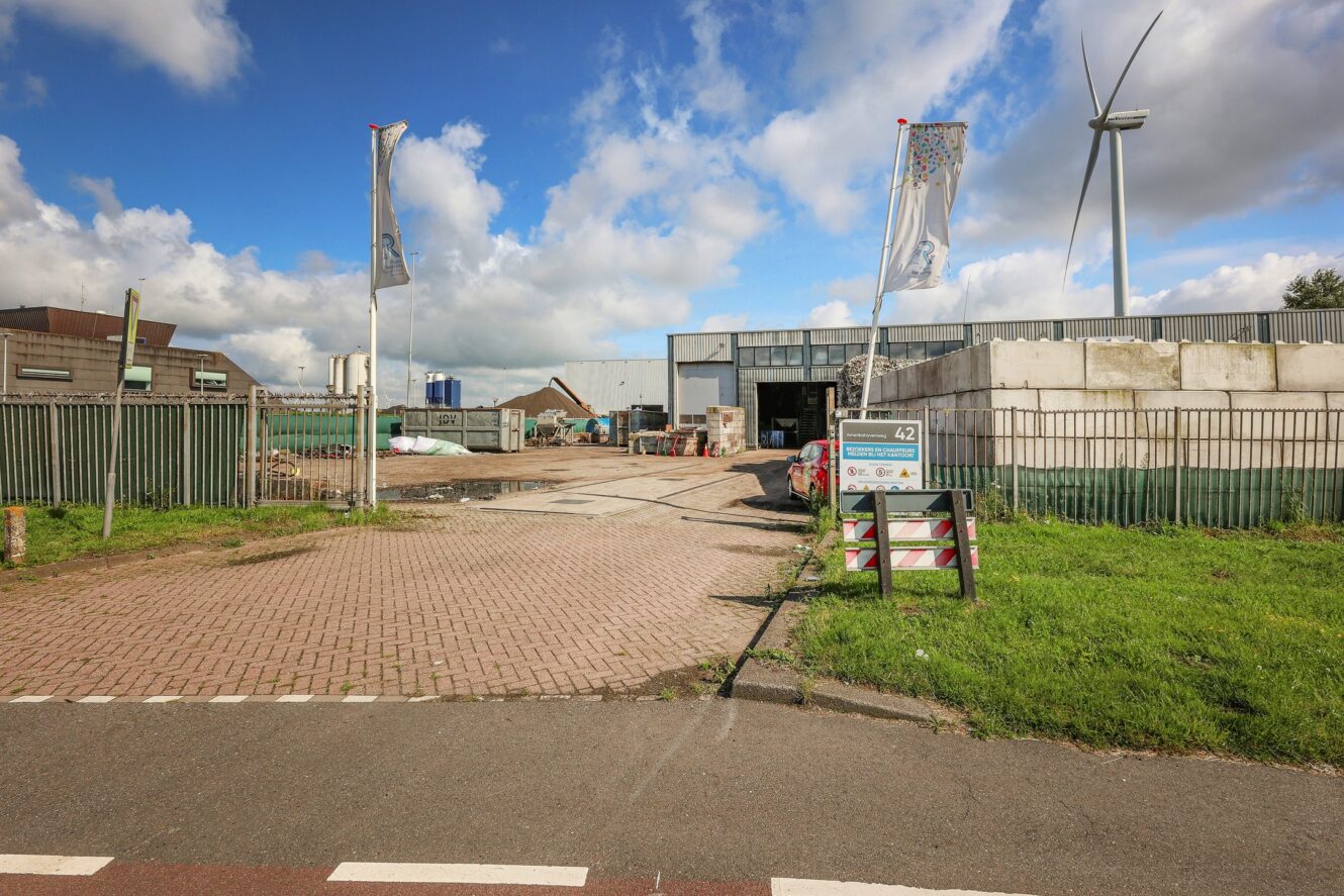 Afgesloten ingang van een industrieterrein met grote magazijngebouwen, bouwmaterialen, twee vlaggenmasten, een uithangbord en een windturbine op de achtergrond onder een gedeeltelijk bewolkte hemel.