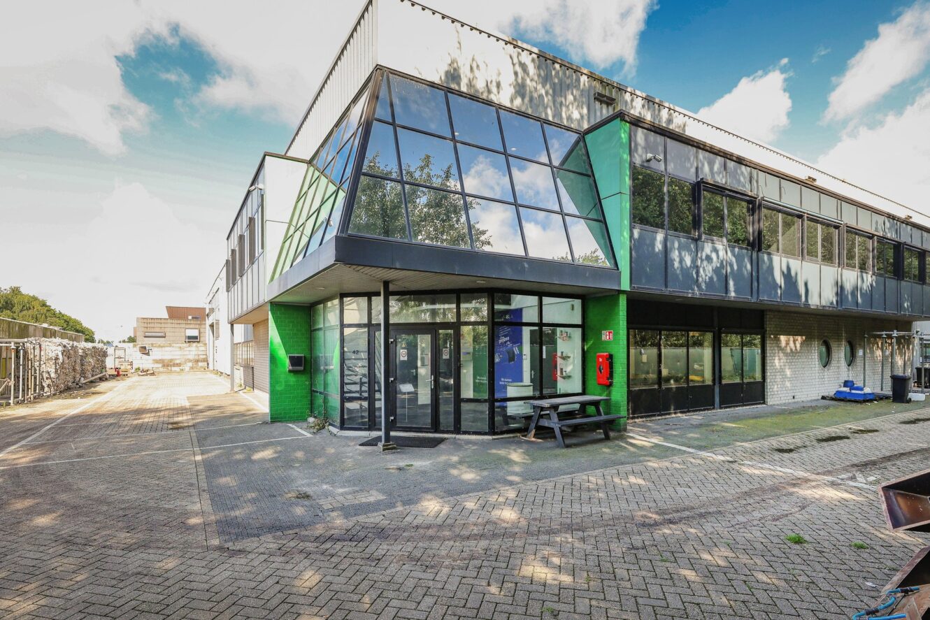 Modern commercieel of industrieel gebouw van twee verdiepingen met grote glazen ramen, groene accenten en een geplaveide binnenplaats. Een picknicktafel en brandblusser staan bij de ingang.
