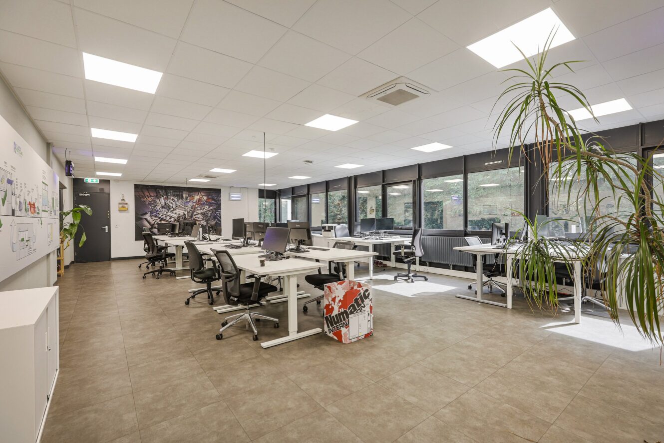 Modern open kantoor met meerdere bureaus, stoelen, computers, grote ramen, heldere verlichting en kamerplanten. De ruimte ziet er schoon, georganiseerd en zonder mensen uit.