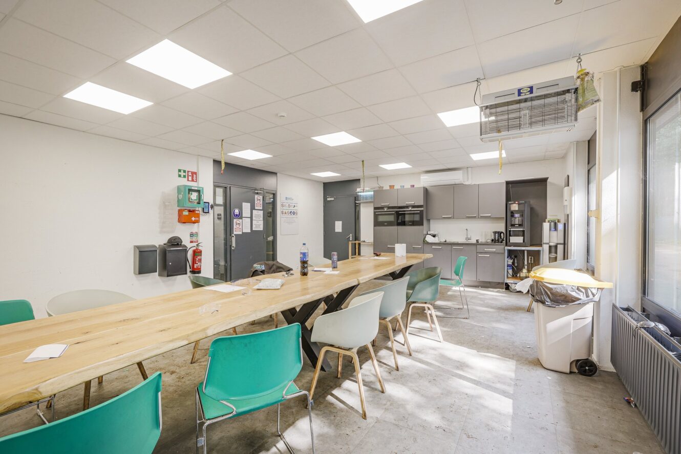 Een moderne pauzeruimte met een lange houten tafel, groene en witte stoelen, een keukentje, apparaten, prullenbakken en tl-verlichting boven het hoofd.