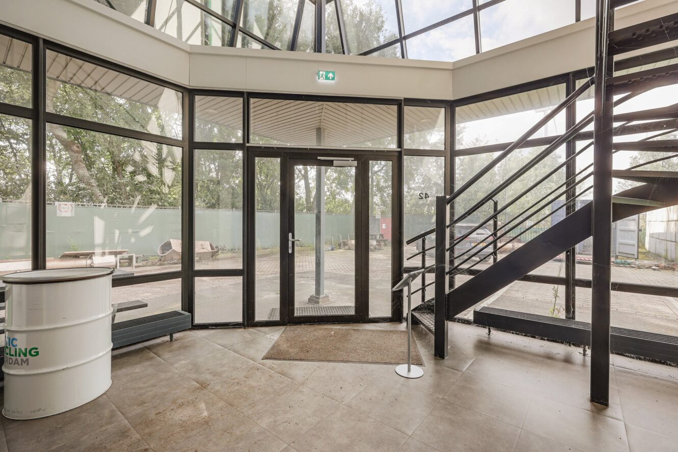 Een glazen entree met een zwart omlijste deur, metalen trap, tegelvloer en trommelvormige tafel, die leidt naar een buitenruimte met bouwmaterialen.