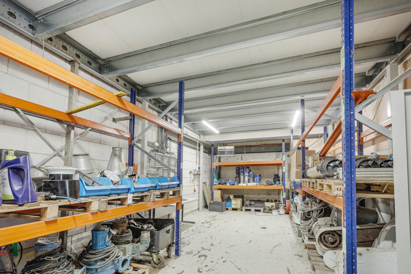 Industriële opslagruimte met metalen rekken, diverse gereedschappen, reserveonderdelen en machineonderdelen georganiseerd op rekken en pallets op een betonnen vloer.