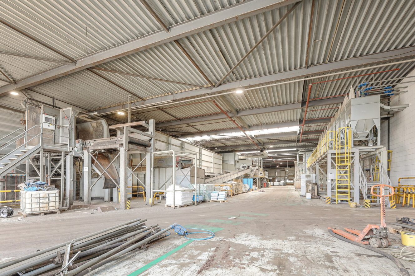 Industrieel magazijninterieur met metalen constructies, machines, trappen en verspreide apparatuur. De ruimte is ruim, goed verlicht en lijkt te worden gebruikt voor materiaalverwerking of opslag.