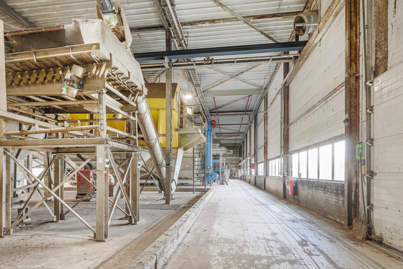 Industrieel interieur met grote metalen machines aan de linkerkant, hoog plafond, zichtbare balken en een brede lege gang met ramen aan de rechterkant.