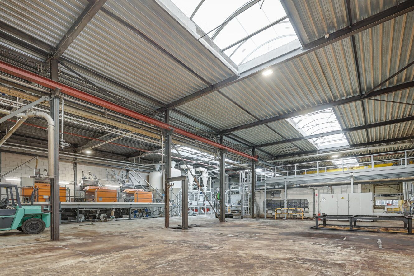 Industrieel magazijninterieur met hoge plafonds, metalen balken, dakramen en grote machines langs de muren. Aan de linkerkant staat een groene vorkheftruck geparkeerd.