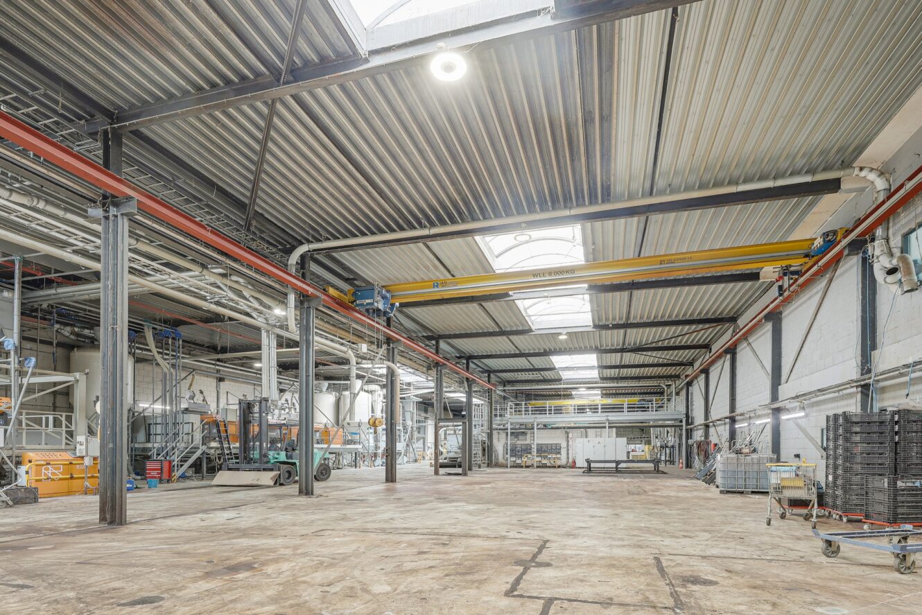 Groot, leeg industrieel pakhuis met hoge plafonds, dakramen, zichtbare leidingen en verspreide apparatuur en pallets langs de muren.