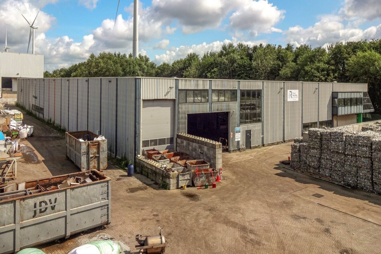 Industriële recyclingfaciliteit met groot metalen gebouw, stapels gesorteerde metalen materialen, afvalcontainers en windturbines op de achtergrond onder een gedeeltelijk bewolkte hemel.